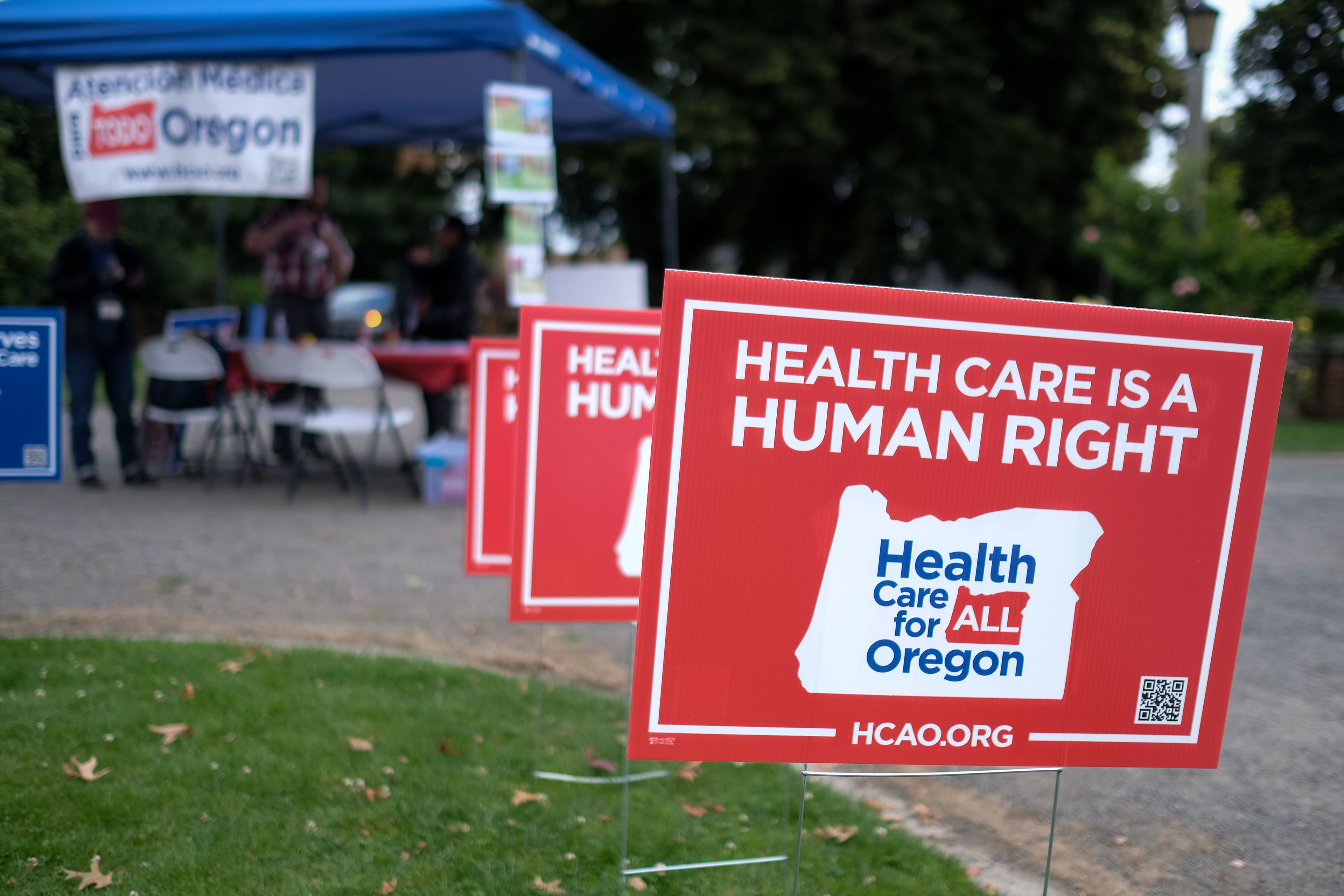 Plastic lawn signs that say "Health Care Is A Human Right" line a sidewalk. In the background, a tent with information about Oregon Healthcare For All is visible.