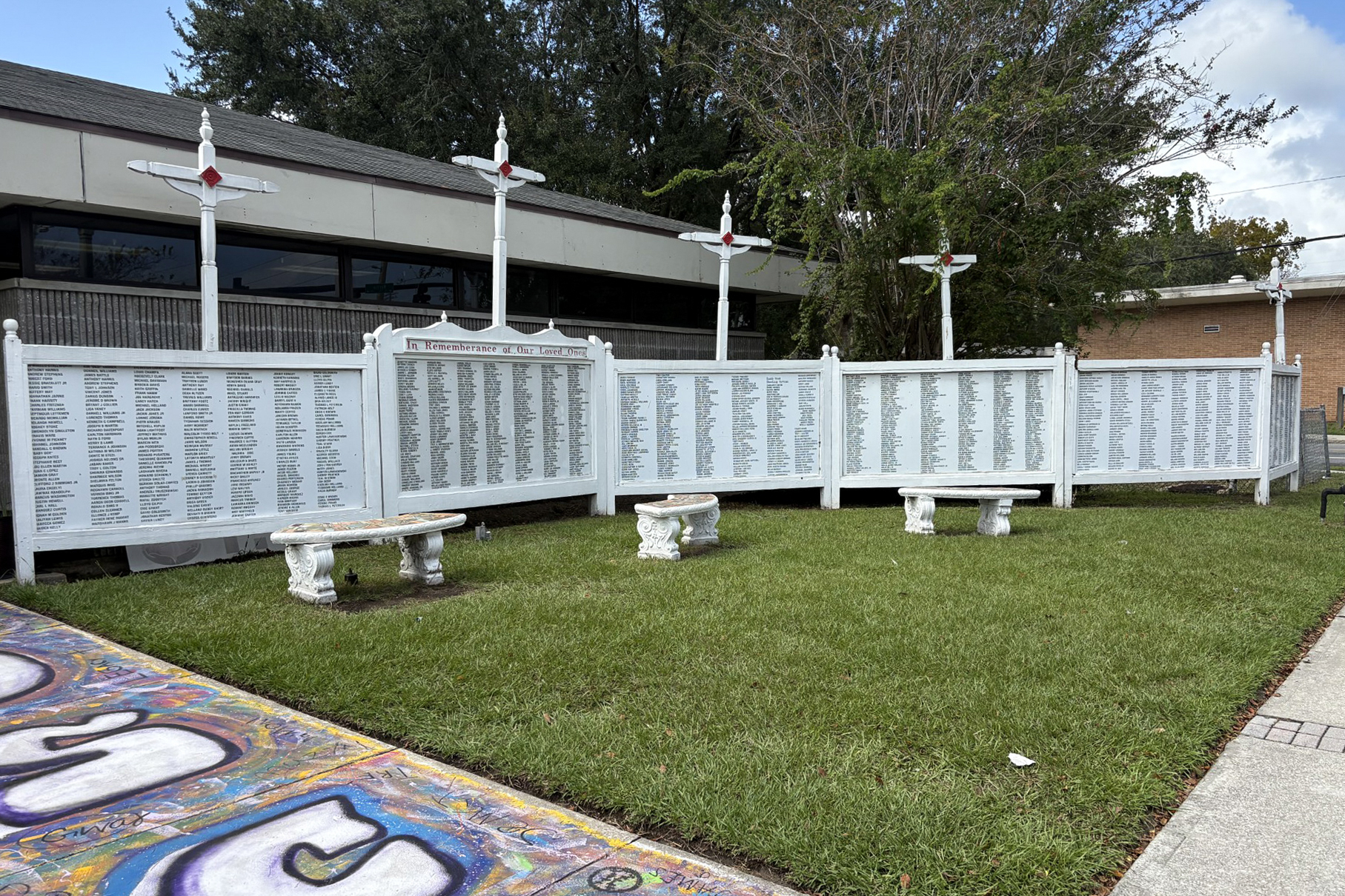 A photo of a white memorial wall with benches in front of it. Writing on it reads, "In rememberance of our loved ones," and lists the names of those killed by gun violence in the community. Four crosses adorn the top of the wall. 