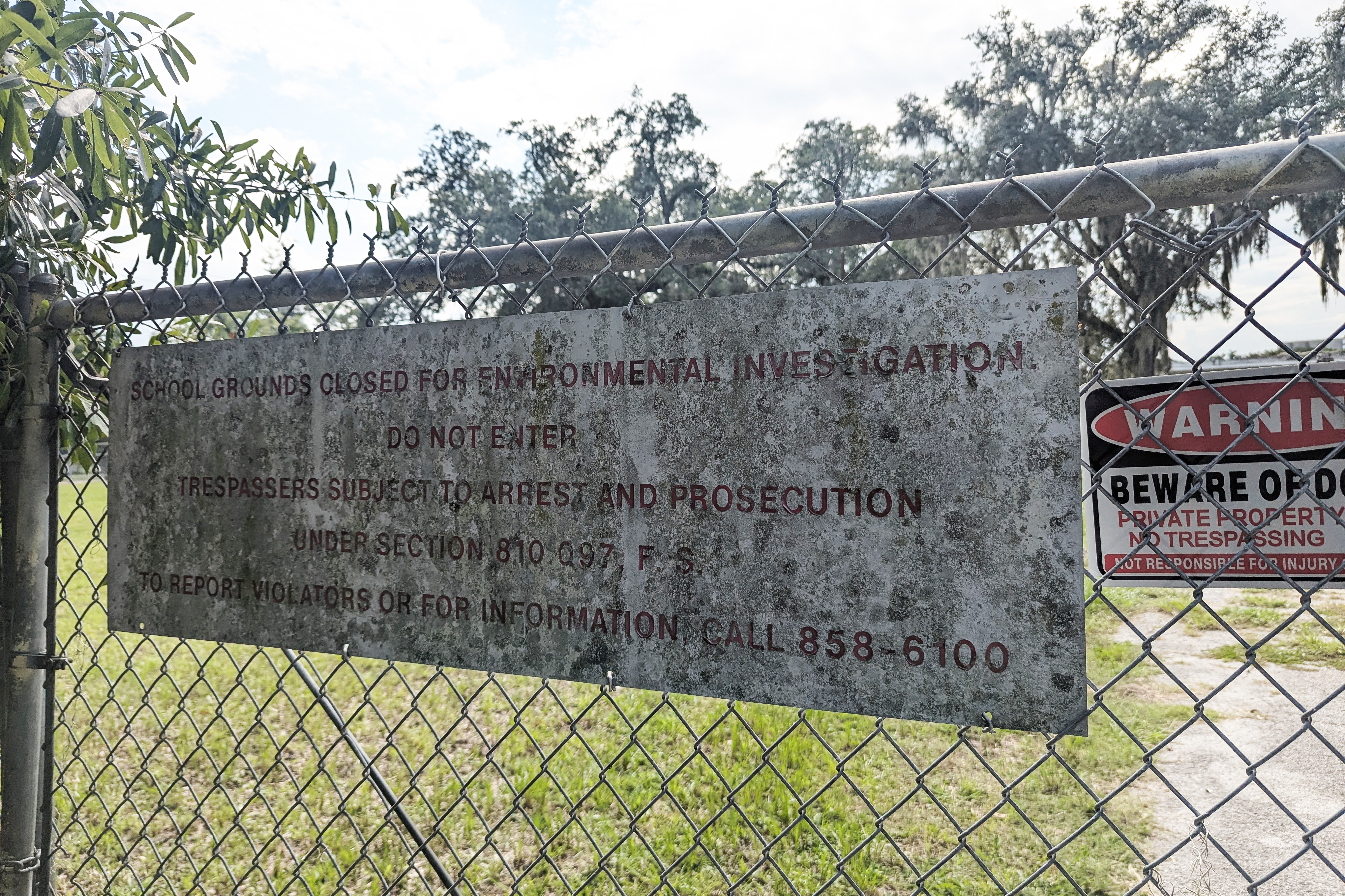 A dirty sign on a chain-link fence in front of a school reads, "School grounds closed for environmental investigation. Do not enter. Trespassers subject to arrest and prosecution. Under Section 8.10.097.F.S. To report violators or for information, call 858-6100."