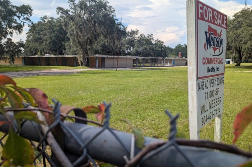 A photo shows a boarded-up school in the background. A sign in the midground advertises the property for sale. A chain-link fence is seen blurred in the foreground.