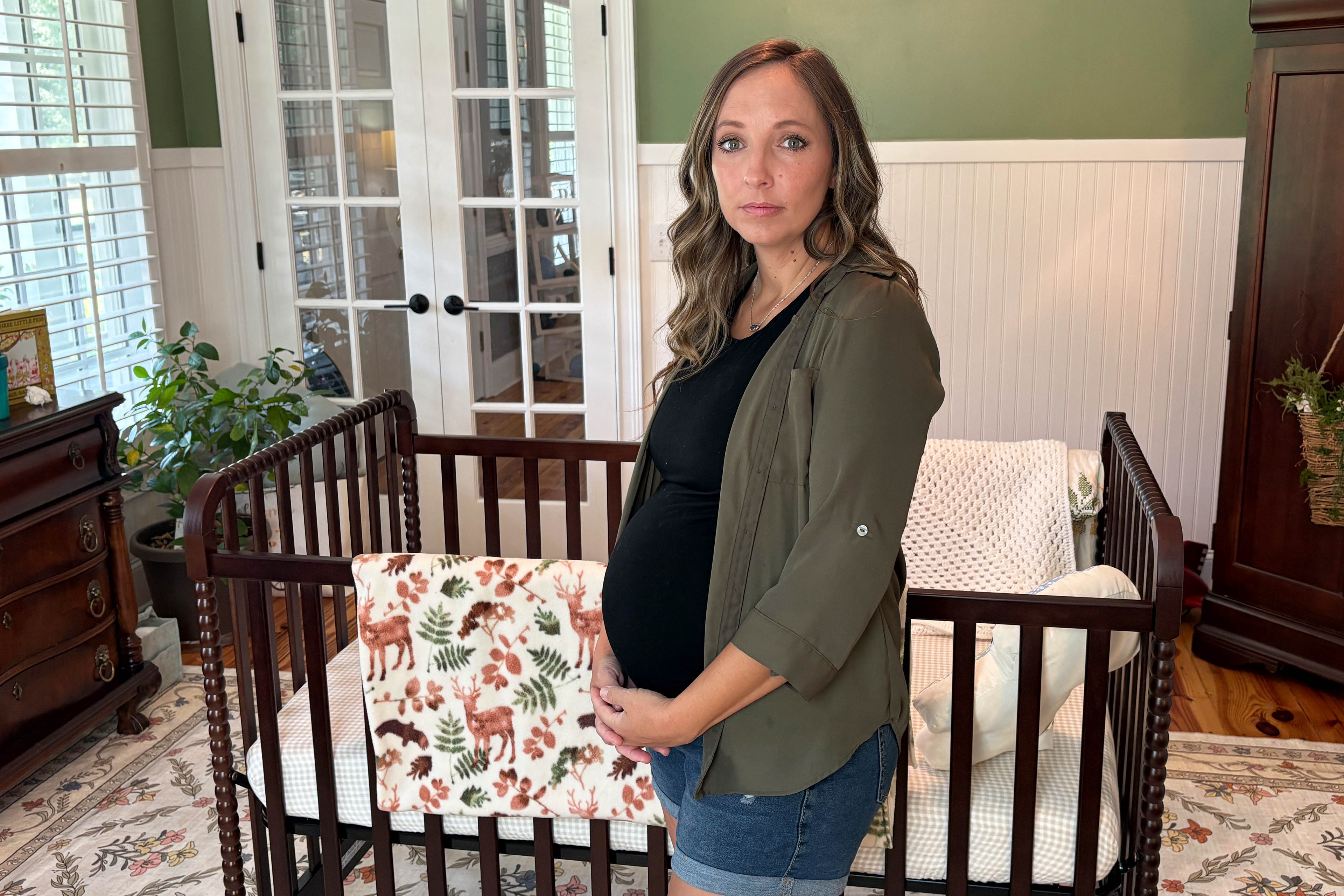 A pregnant woman, resting her hands on her lower stomach, stands beside a crib for a baby in her home.