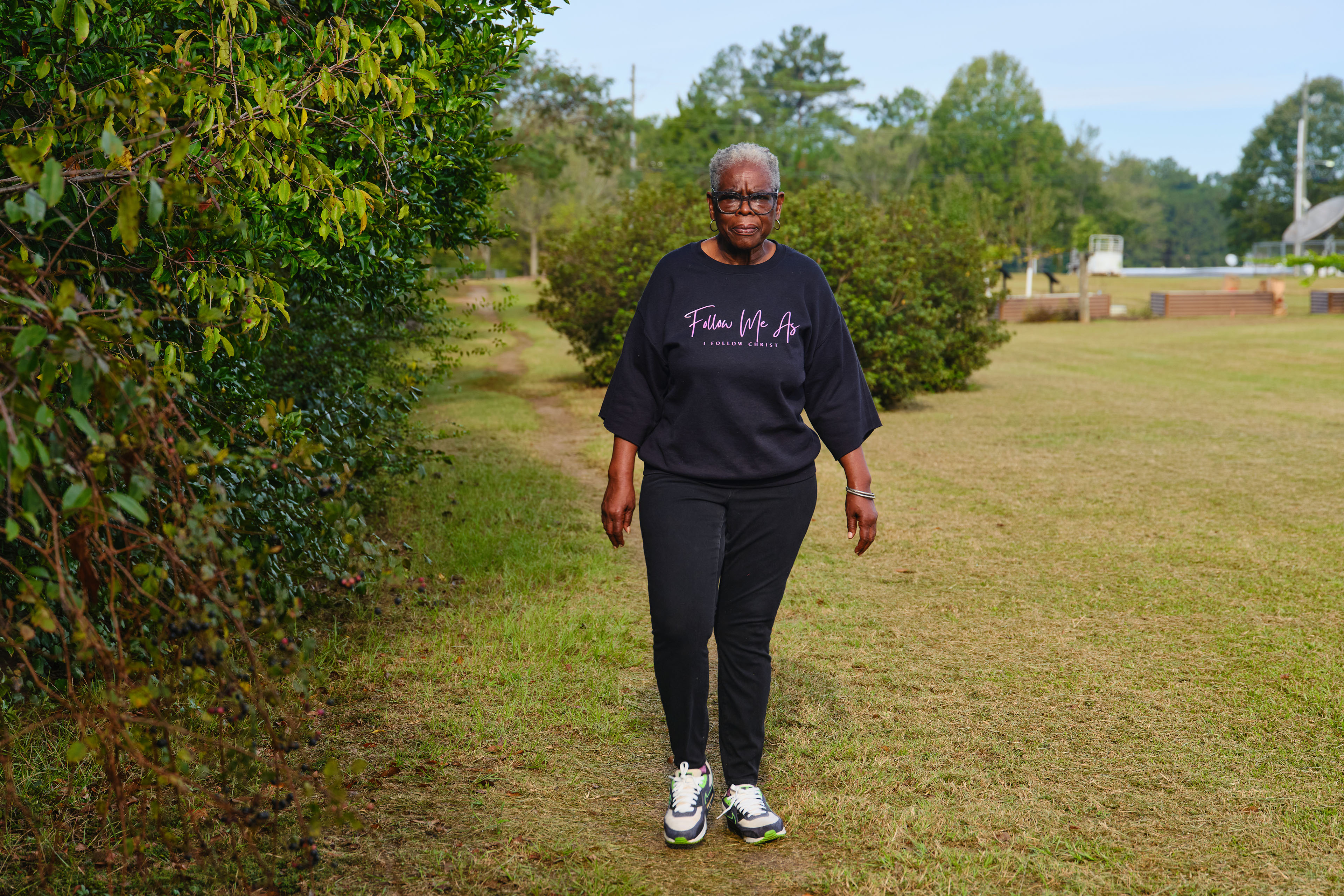 A woman wearing glasses and a black sweatshirt walks along a trail next to a field