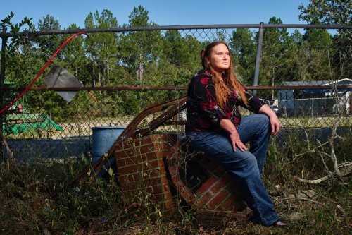A photo of a woman sitting on remains of a brick oven. A chain link fence is seen behind her. The general area is overgrown.
