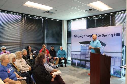 A man stands near to a podium. Behind him is a large sign that reads, "Bringing a hospital to Spring Hill." A group of people are seated in front of him, listening.