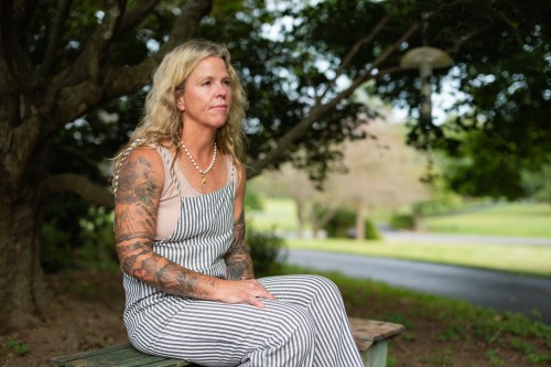 A photo of a woman seated on a bench in the shade of a tree.