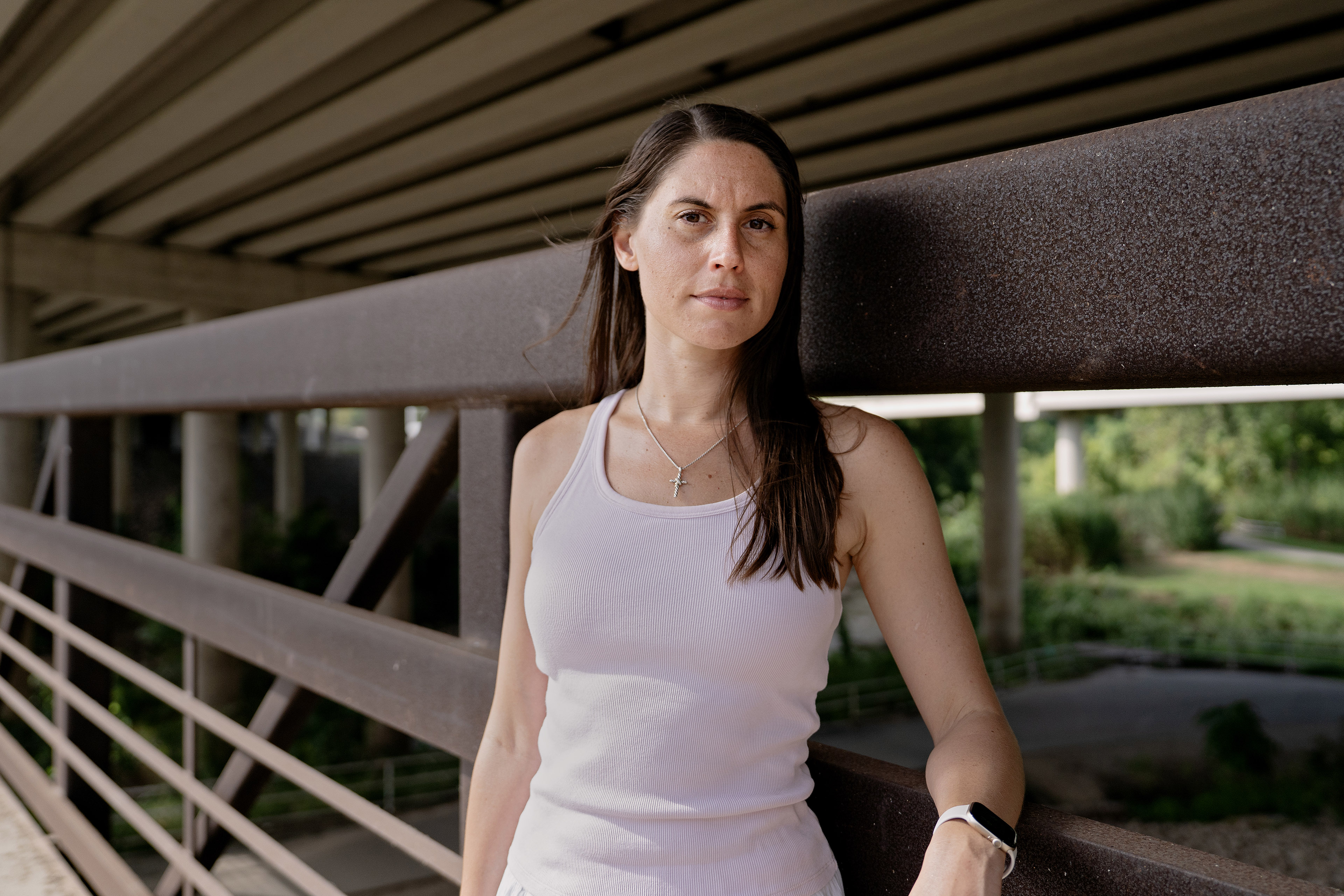 A woman with brown hair, wearing a white tank top, stands on a bridge